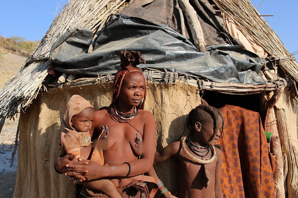 一位妇女抱着孩子站在屋前，在辛巴部落中，女人通常要承担更大的家庭负担。