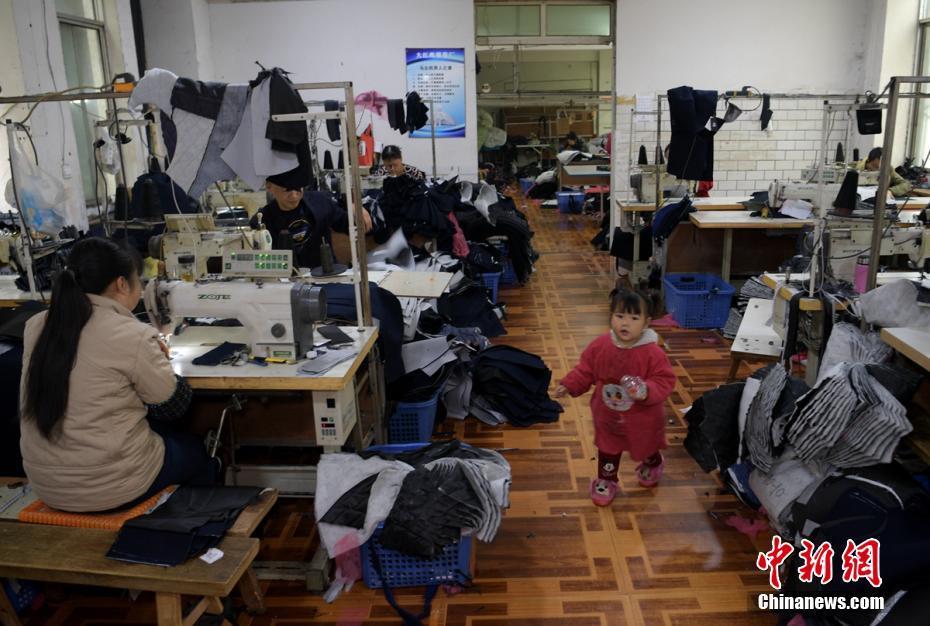 成都郫都区安靖镇土地村某服装加工厂车间内一小女孩正在玩耍，其母亲正坐在缝纫机前赶制服装。